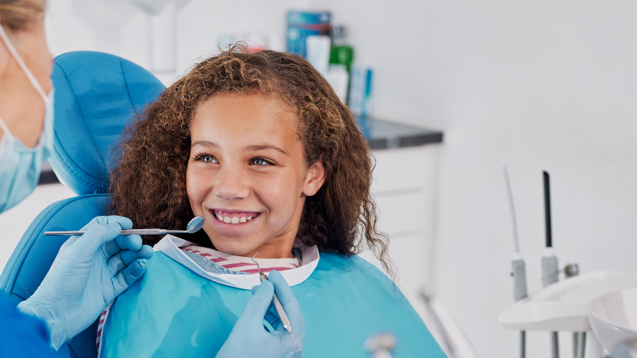 a child sits in a dental chair with a smile on her face while a dentist examines her teeth