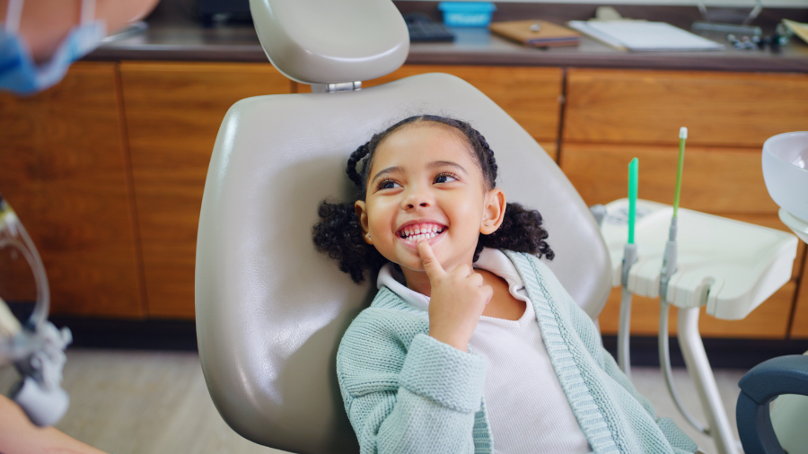 a child sits in the dental chair and points to her teeth with a smile